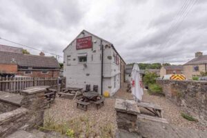 Property for sale Outdoor seating area with wooden benches and tables beside a stone building marked "Petton Arms." This detached public house for sale in Burnopfield, Newcastle upon Tyne offers a freehold property with beer garden, surrounded by stone walls and houses. Presented by Palace Auctions