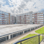 Property for sale A wide view of a large, rectangular apartment complex with several stories, perfect for a buy-to-let West Bridgford investment. In the foreground are a paved walkway and green grass, all beneath a cloudy sky. Presented by Palace Auctions
