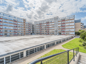Property for sale A wide view of a large, rectangular apartment complex with several stories, perfect for a buy-to-let West Bridgford investment. In the foreground are a paved walkway and green grass, all beneath a cloudy sky. Presented by Palace Auctions