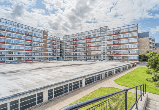 Property for sale A wide view of a large, rectangular apartment complex with several stories, perfect for a buy-to-let West Bridgford investment. In the foreground are a paved walkway and green grass, all beneath a cloudy sky. Presented by Palace Auctions
