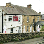 Property for sale A stone and white-painted building labeled "Brook House" stands behind a stone wall on a sloped street—a great example of a traditional public house conversion in the Newcastle area. Outdoor umbrellas and a parked car sit at its entrance. Presented by Palace Auctions