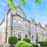 Property for sale A large, light-brick Victorian-style house with arched windows, bay windows, and neatly trimmed green bushes in front—ideal for a WA14 residential investment—set against a bright blue sky with leafy branches overhead. Presented by Palace Auctions