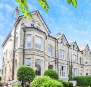 Property for sale A large, light-brick Victorian-style house with arched windows, bay windows, and neatly trimmed green bushes in front—ideal for a WA14 residential investment—set against a bright blue sky with leafy branches overhead. Presented by Palace Auctions