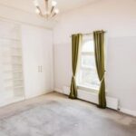 Property for sale A bright, empty room in this Altrincham period flat features light gray carpet, a window with green curtains, built-in white shelves and closet, plus a modern ceiling light fixture. Presented by Palace Auctions