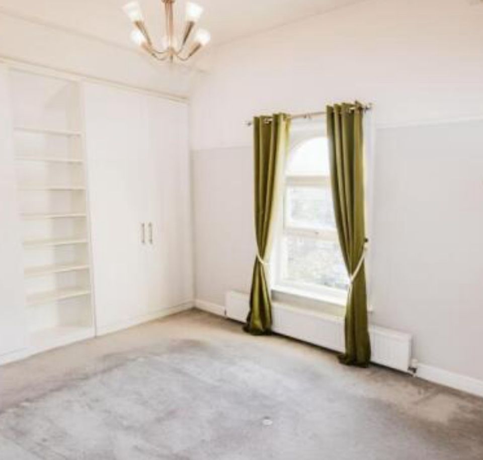 Property for sale A bright, empty room in this Altrincham period flat features light gray carpet, a window with green curtains, built-in white shelves and closet, plus a modern ceiling light fixture. Presented by Palace Auctions