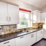 Property for sale A modern kitchen in this Bowdon 4-bed flat features white cabinets, a black countertop, tiled backsplash, stainless steel sink, and a window with a red blind letting in natural light. Presented by Palace Auctions