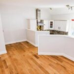 Property for sale A bright, unfurnished room with light wooden flooring and white walls in this Heald Road apartment auction, featuring a partial view of a modern kitchen with white cabinets, tiled backsplash, and a stainless steel range hood. Presented by Palace Auctions