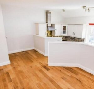 Property for sale A bright, unfurnished room with light wooden flooring and white walls in this Heald Road apartment auction, featuring a partial view of a modern kitchen with white cabinets, tiled backsplash, and a stainless steel range hood. Presented by Palace Auctions