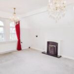 Property for sale A bright, empty room in this Bowdon 4-bed flat features two crystal chandeliers, a fireplace with a white mantel, light grey carpet, white walls, and large windows with red curtains allowing natural light to enter. Presented by Palace Auctions