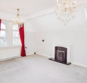 Property for sale A bright, empty room in this Bowdon 4-bed flat features two crystal chandeliers, a fireplace with a white mantel, light grey carpet, white walls, and large windows with red curtains allowing natural light to enter. Presented by Palace Auctions