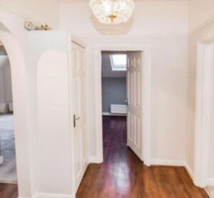 Property for sale A bright hallway in this Altrincham period flat boasts white walls, wood flooring, and a chandelier. Two doors reveal adjoining rooms—one with a skylight and radiator, another with a partially visible carpeted space. Presented by Palace Auctions