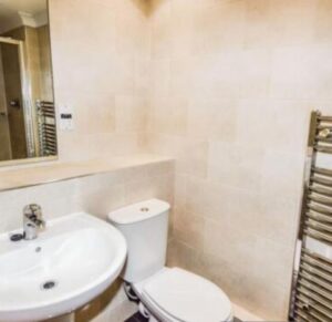 Property for sale A modern bathroom in this Altrincham period flat features a white sink, toilet, wall-mounted mirror, beige tiled walls, and a metallic heated towel rail on the right. Presented by Palace Auctions