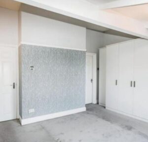 Property for sale A bright, empty room with light gray carpet, white walls, a blue patterned accent wall, two white doors, and a large white wardrobe with black handles—ideal for those seeking a WA14 residential investment on Heald Road or Bowdon investment property. Presented by Palace Auctions