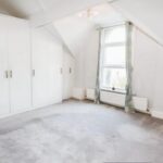 Property for sale A bright, empty attic room in an Altrincham period flat, featuring white walls, built-in wardrobes, a large arched window with light green curtains, grey carpet, and a radiator beneath the window. Presented by Palace Auctions