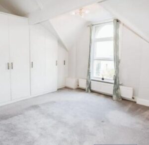 Property for sale A bright, empty attic room in an Altrincham period flat, featuring white walls, built-in wardrobes, a large arched window with light green curtains, grey carpet, and a radiator beneath the window. Presented by Palace Auctions