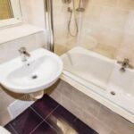 Property for sale A small bathroom in a Bowdon investment property features a white wall-mounted sink, rectangular bathtub with showerhead, beige tiled walls, dark brown flooring, and a mirror above the sink—ideal for WA14 residential investment. Presented by Palace Auctions
