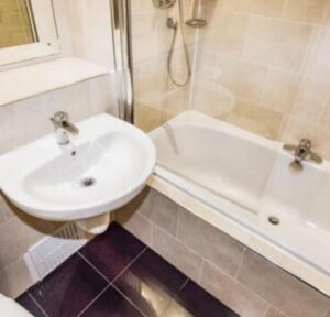 Property for sale A small bathroom in a Bowdon investment property features a white wall-mounted sink, rectangular bathtub with showerhead, beige tiled walls, dark brown flooring, and a mirror above the sink—ideal for WA14 residential investment. Presented by Palace Auctions