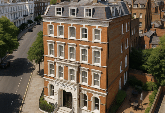 Property for sale A four-story red-brick Boutique Hotel Chelsea with white trim, named "The Overley Hotel," stands on a street corner surrounded by greenery, outdoor seating, and classic townhouses under a clear blue sky. Presented by Palace Auctions