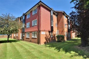 Property for sale A three-story red brick apartment building with white-framed windows, surrounded by neatly mowed grass and trees under a clear blue sky. Ideal for first-time buyer flats Staines-upon-Thames, a sign on the building reads "RENALD COURT. Presented by Palace Auctions