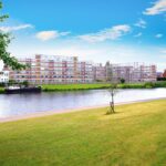 Property for sale A riverside scene with green grass, a few scattered trees, and a bench in the foreground. Across the river, a modern apartment building—ideal for buy-to-let West Bridgford investors—stands beside older homes under a bright blue sky with clouds. Presented by Palace Auctions