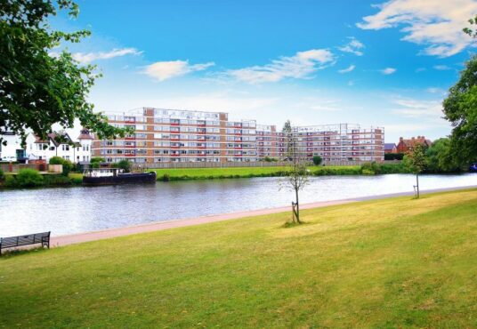 Property for sale A riverside scene with green grass, a few scattered trees, and a bench in the foreground. Across the river, a modern apartment building—ideal for buy-to-let West Bridgford investors—stands beside older homes under a bright blue sky with clouds. Presented by Palace Auctions
