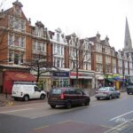 Property for sale A row of Victorian-style buildings with shops at street level lines busy 2C Hamilton Road W5 2EQ, where parked and moving cars pass leafless trees along the sidewalk, and a church spire rises in the background. Presented by Palace Auctions