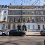 Property for sale A row of Grade II* listed townhouses with brick façades and white lower levels, lined with black railings. Several parked cars are on the street in front, and bare tree branches are visible above this historic London property investment. Presented by Palace Auctions
