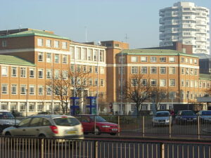Property for sale Cars drive past a large, multi-story brick building with green roofs near the Orchard Rise Croydon development land. In the background, a tall, white modern apartment tower stands, with leafless trees lining the street in front. Presented by Palace Auctions