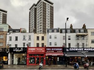 Property for sale A row of restaurants and shops with colorful signs lines this city street, set against tall multi-unit rental property buildings in the background, with several parked scooters out front on a cloudy day. Presented by Palace Auctions
