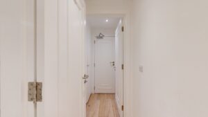 Property for sale A narrow hallway with white walls and doors, light wood flooring, and a glimpse of a ceiling light at the far end near a closed door—ideal for a high yield London property or Edgware Road property investment. Presented by Palace Auctions