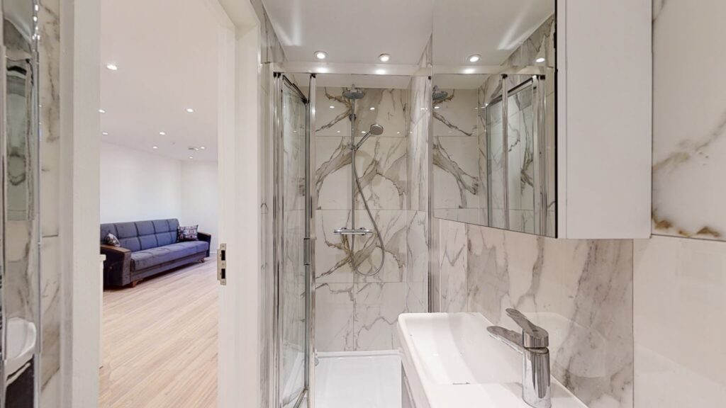 Property for sale Modern bathroom with marble-patterned walls, glass shower enclosure, dual showerheads, and mirrored cabinets—ideal for a high yield London property. A cozy living area with a sofa is visible in the background, perfect for multi-unit rental property appeal. Presented by Palace Auctions