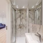 Property for sale Modern bathroom with marble-patterned walls, glass shower enclosure, dual showerheads, and mirrored cabinets—ideal for a high yield London property. A cozy living area with a sofa is visible in the background, perfect for multi-unit rental property appeal. Presented by Palace Auctions