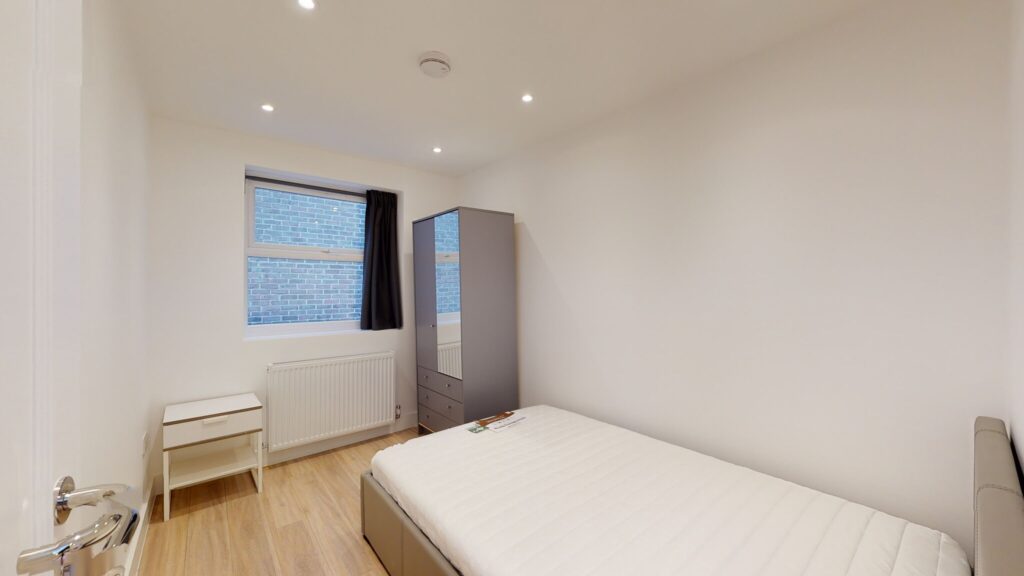 Property for sale A simple bedroom with white walls, a single bed with a white mattress, a desk and chair by a window with a dark curtain, a tall mirrored wardrobe, and wood flooring—ideal for those seeking high yield London property. Presented by Palace Auctions