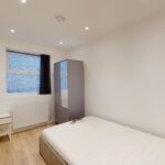 Property for sale A simple bedroom with white walls, a single bed with a white mattress, a desk and chair by a window with a dark curtain, a tall mirrored wardrobe, and wood flooring—ideal for those seeking high yield London property. Presented by Palace Auctions