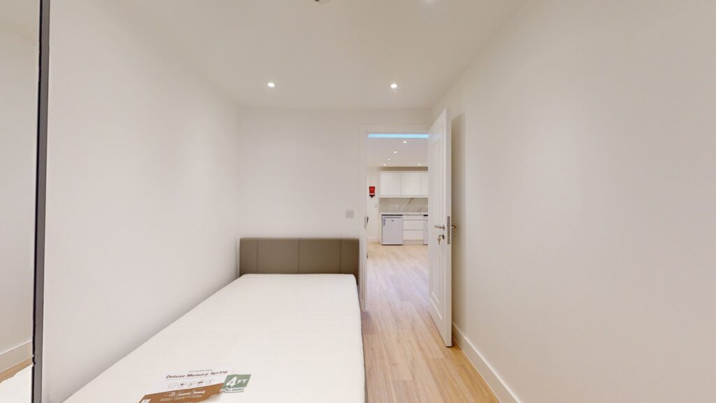 Property for sale A small, minimal bedroom with a single bed, white walls, and wood flooring—part of a mixed-use freehold London property—features an open door leading to a bright kitchen area with modern appliances. Presented by Palace Auctions