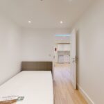Property for sale A small, minimal bedroom with a single bed, white walls, and wood flooring—part of a mixed-use freehold London property—features an open door leading to a bright kitchen area with modern appliances. Presented by Palace Auctions