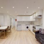 Property for sale Modern open-plan kitchen and living area with white cabinets, built-in appliances, a dining table for six, and a dark sofa on light wooden floors—ideal for an Edgware Road property investment or multi-unit rental property. Presented by Palace Auctions