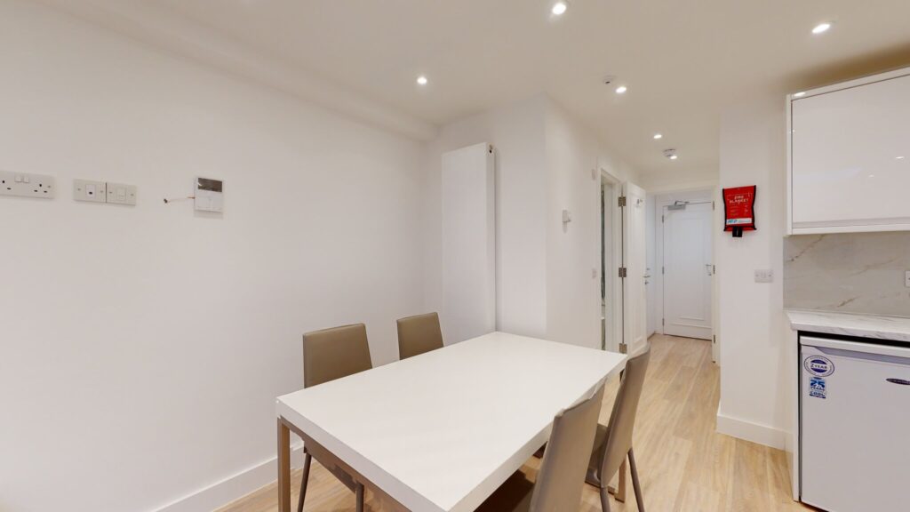 Property for sale A modern, minimalistic dining area with a white table and four beige chairs, set within a high yield London property. Light wood flooring and white walls create an airy space that connects to a hallway and partially visible kitchen appliances. Presented by Palace Auctions