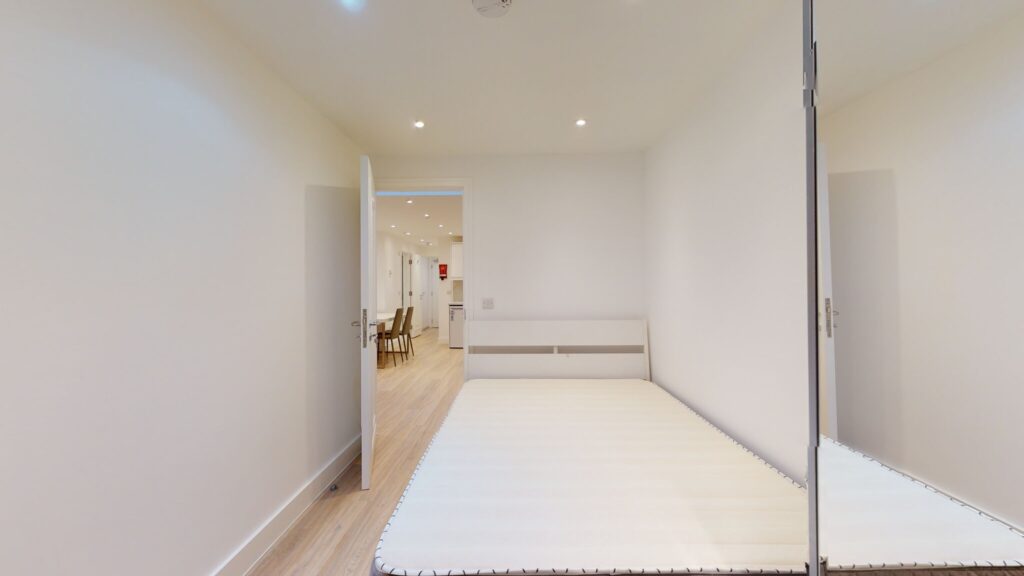 Property for sale A minimal bedroom with a white bed frame and no mattress, light wooden floor, and white walls—ideal for a high yield London property—with an open door revealing a hallway, dining chairs, and kitchen area in this multi-unit rental property. Presented by Palace Auctions