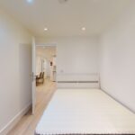 Property for sale A minimal bedroom with a white bed frame and no mattress, light wooden floor, and white walls—ideal for a high yield London property—with an open door revealing a hallway, dining chairs, and kitchen area in this multi-unit rental property. Presented by Palace Auctions