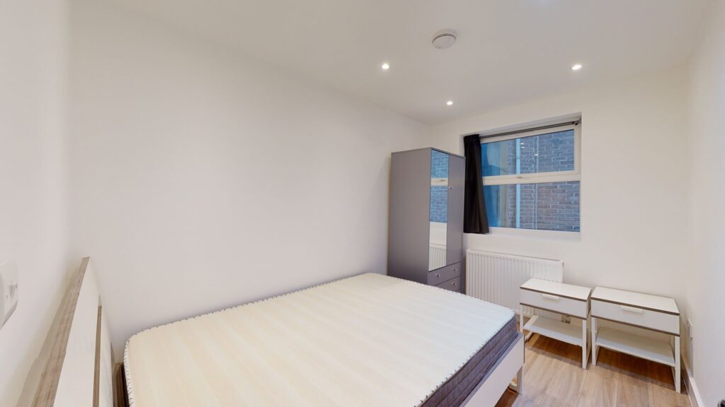 Property for sale A small, modern bedroom in a multi-unit rental property features a double bed, two white nightstands, a mirrored wardrobe, wooden flooring, black-curtained window, and white walls lit by ceiling lights. Presented by Palace Auctions