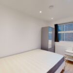 Property for sale A small, modern bedroom in a multi-unit rental property features a double bed, two white nightstands, a mirrored wardrobe, wooden flooring, black-curtained window, and white walls lit by ceiling lights. Presented by Palace Auctions