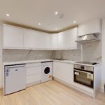 Property for sale Modern kitchen with white cabinets, marble backsplash, small fridge, and electric stove—ideal for a high yield London property. Light wood floors and a glimpse of the gray sofa enhance this inviting multi-unit rental property setting. Presented by Palace Auctions