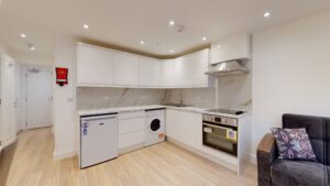 Property for sale Modern kitchen with white cabinets, marble backsplash, small fridge, and electric stove—ideal for a high yield London property. Light wood floors and a glimpse of the gray sofa enhance this inviting multi-unit rental property setting. Presented by Palace Auctions