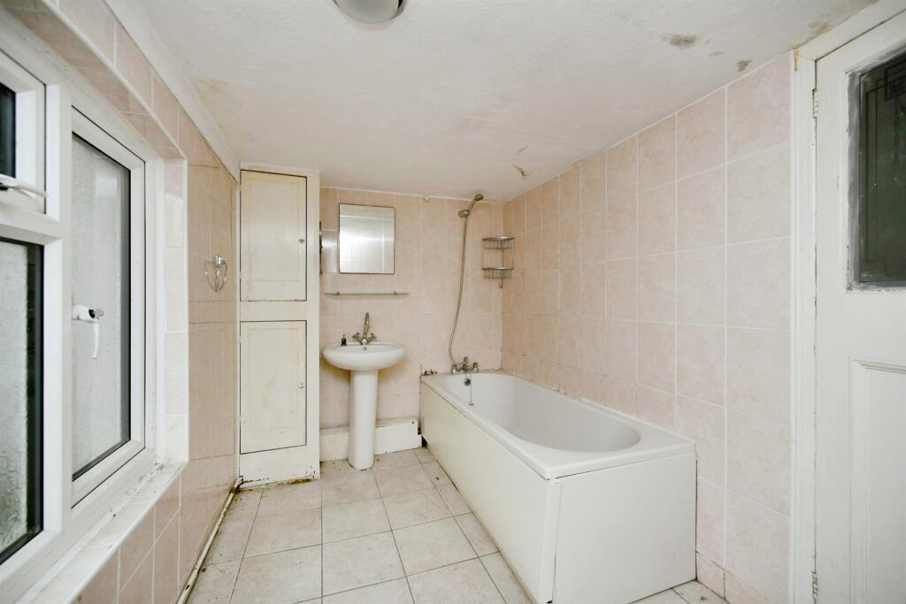 Property for sale A narrow bathroom in a 2-bed maisonette Newhaven, with light pink tiles, a white bathtub and shower, pedestal sink, wall cabinet, small mirror, and frosted window. The room appears old with visible signs of wear—potential South Road investment. Presented by Palace Auctions
