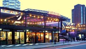 Property for sale Boxpark, an urban food and retail complex built from shipping containers and lit by string lights at dusk, sits near the Orchard Rise Croydon development land. People stroll nearby as a tall building stands in the background. Presented by Palace Auctions