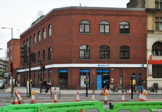Property for sale A red-brick building on a city street corner houses a Barclays bank branch. Cyclists and pedestrians pass by, with green construction barriers and traffic lights framing this Whitechapel Road mixed-use development opportunity. Presented by Palace Auctions