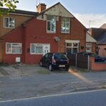 Property for sale A two-story semi-detached house with a red and beige exterior, ideal as an Abbey Lane Investment Property, features a black car in the driveway and a brick wall separating it from neighbors on a quiet suburban street. Presented by Palace Auctions