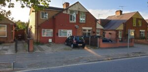 Property for sale A two-story semi-detached house with a red and beige exterior, ideal as an Abbey Lane Investment Property, features a black car in the driveway and a brick wall separating it from neighbors on a quiet suburban street. Presented by Palace Auctions