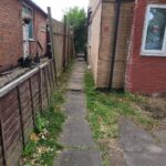 Property for sale A narrow concrete path runs between two brick buildings, with grass and plants along the edges—a potential investment property in Leicester. One building has a window, the other potted plants, and a wooden fence divides this split unit investment area. Presented by Palace Auctions